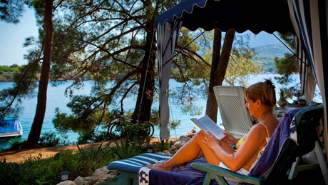 A person relaxing and reading by the seaside under a shaded canopy with trees and water in the background