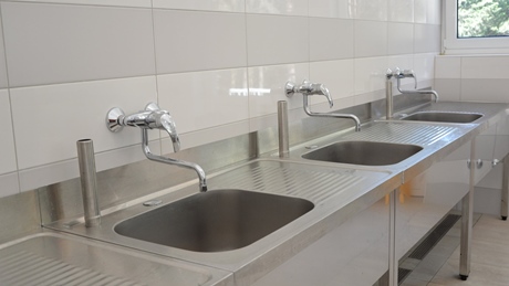 A row of stainless steel sinks with faucets in a tiled room